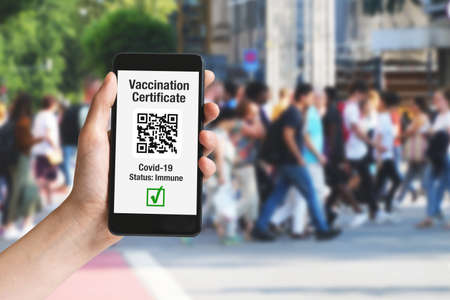 Hand holding mobile phone in city with vaccination certificate on display as symbol for immunization witth people in backgroundの写真素材
