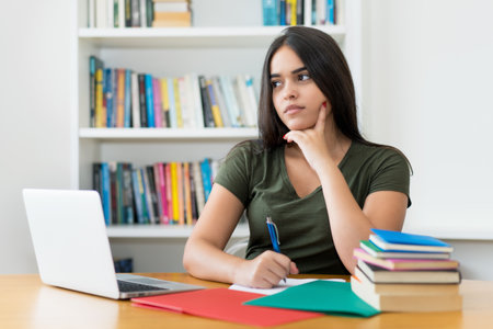Thinking spanish female student at desk indoors at libraryの写真素材