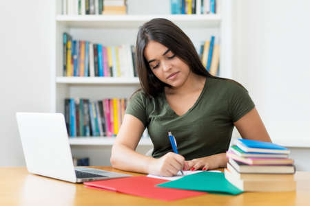 Young spanish female student writing notes at desk indoors at libraryの写真素材
