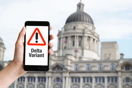Hand holding mobile phone with with warning message delta variant of coronavirus and historic building at Liverpoolの写真素材