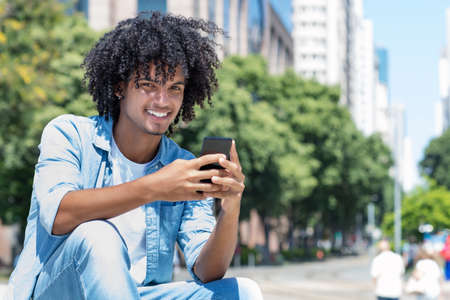 Laughing latin american young adult man sending message with mobile phone outdoor in city in summerの写真素材
