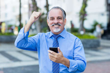 Cheering latin american senior businessman receiving good news on phone outdoor in city in summerの写真素材