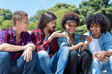 Group of latin and african american young adults watching clip on phone outdoor in city in summerの写真素材