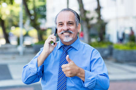 Happy mexican senior businessman at phone showing thumb up outdoor in city in summerの写真素材