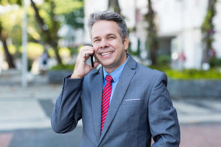 Laughing mature businessman at phone with suit and gray hair outdoor in city in summerの写真素材