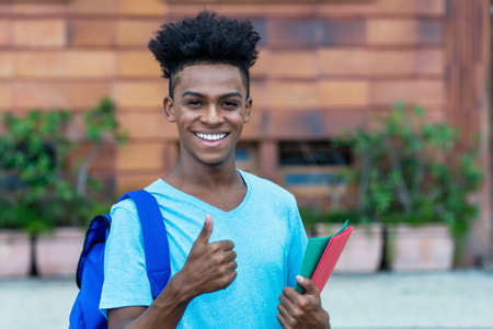 Successful afro american male student with backpack and paperwork showing thumb up outdoor in city in summerの写真素材