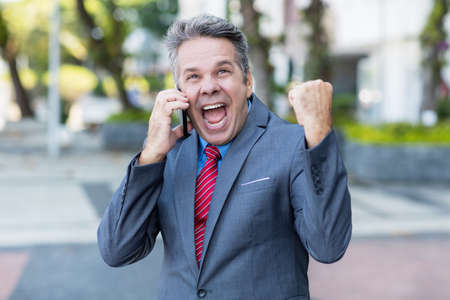 Successful mature businessman at phone with suit and gray hair outdoor in city in summerの写真素材