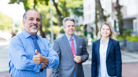 Smart mexican senior businessman with colleagues showing thumb up outdoor in city in summerの写真素材