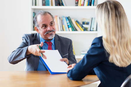 Hispanic senior businessman showing purchase contract to buyer at desk at officeの写真素材