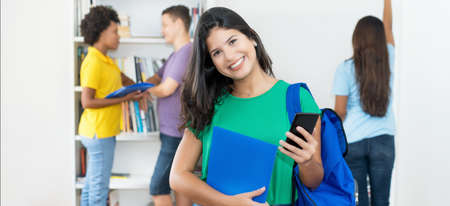 Pretty spanish female student with backpack and mobile phone at classroom of collegeの写真素材
