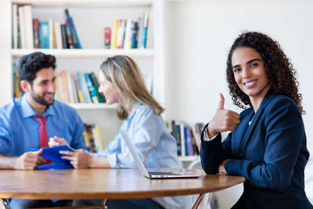 Successful hispanic businesswoman showing thumb up with colleagues at officeの写真素材