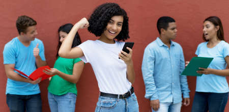 Cheering indian female student receiving good news at phone with group of multi ethnic young adults outdoor in summerの写真素材
