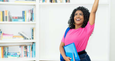 Cheering brazilian female student with backpack and paperwork at classroom of universityの写真素材
