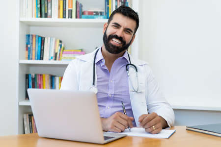 Portrait of happy laughing latin american mature doctor with hipster beard at office of clinicの写真素材