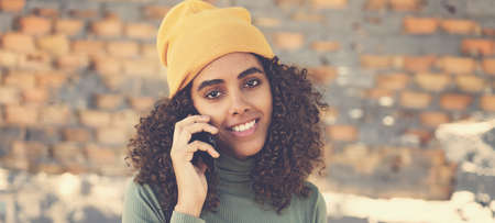 Pretty latin american female young adult with knit hat talking at phone outdoor in urban city styleの写真素材