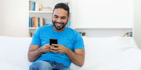 Handsome latin american hipster man with beard sending message at mobile phone indoors at homeの写真素材