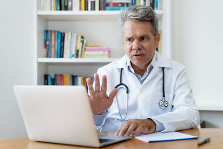 Senior doctor with gray hair gesturing stop to patient at video call at office of hospitalの写真素材