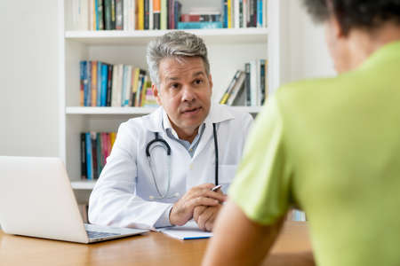 Serious doctor with gray hair talking with patient at office of hospitalの写真素材