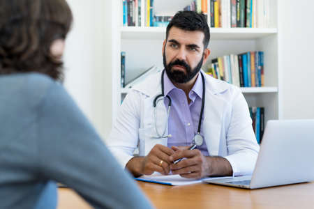 Serious mature doctor with hipster beard listening to patient at office of hospitalの写真素材