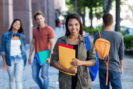 Laughing latin american young adult woman walking in city with group of students outdoors in cityの写真素材