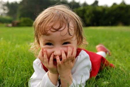 Little laughing girl closing her mouth with hands lying on green grass in the garden summertimeの写真素材