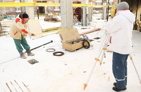 Land surveyor and assistant working with theodolite equipment at a construction site in winter