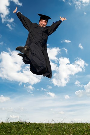 Young smiley graduate student in gown jumping over blue skyの写真素材