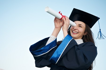 Young laughing graduate girl student in gown looking through diploma outdoorsの写真素材