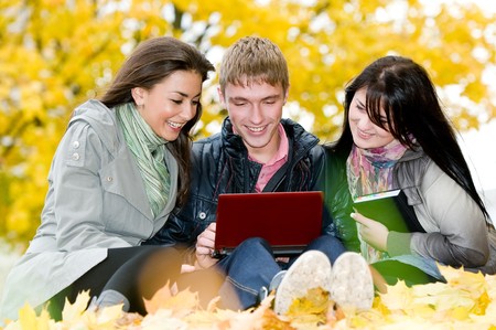 Three students sitting in park on yellow autumn leaves with computer notebookの写真素材