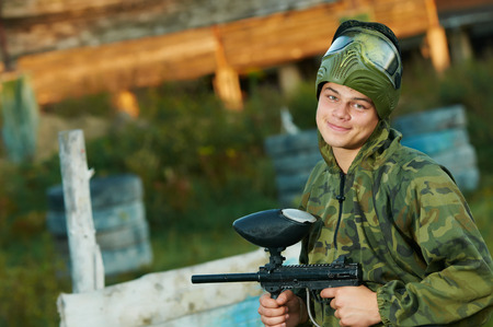 Happy paintball sport player man in protective camouflage uniform and mask with marker gun outdoorsの写真素材