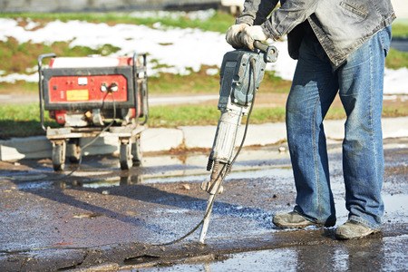 Builder worker with pneumatic hammer drill equipment breaking asphalt at road construction site