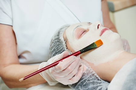 beautician worker with paintbrush applying facial cosmetic mask to female client in beauty salonの写真素材