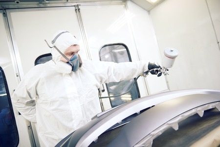 Auto repairman mechanic painter in protective workwear and respirator painting car body bumper in paint chamber at garage service station. Tonedの写真素材