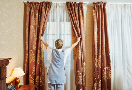 Hotel service. female housekeeping chambermaid worker with opening curtains of window in roomの写真素材