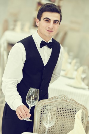 Catering service. Portrait of young male waiter man serving banquet table with knife fork and glass at restaurant or cafeの写真素材