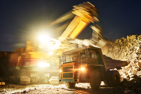 Mining industry. Heavy excavator loading granite rock or iron ore into the huge dump truck at opencast quarry. Duskの写真素材