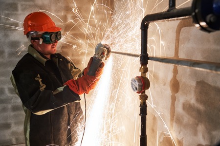 Construction worker in protective glasses cutting metal pipe at building site with sparks by...