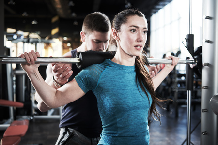 Personal trainer man helps woman work with barbell at squat exercises in gymの写真素材