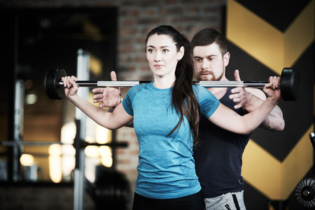 Personal trainer man helps woman work with barbell at squat exercises in gymの写真素材