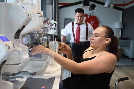 Minsk. Belarus - 06.06.2021: A woman in a wheelchair makes coffee in a coffee machine in a cafe. Behind her is a manager in a white shirt and red tie.のeditorial素材