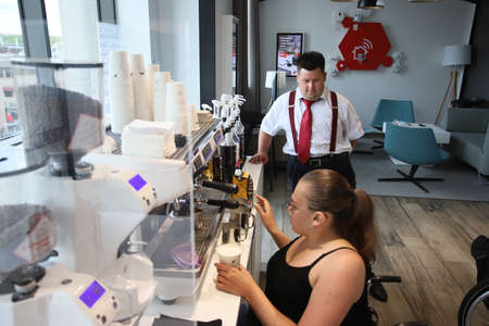 Minsk. Belarus - 06.06.2021: A woman in a wheelchair makes coffee in a coffee machine in a cafe. Behind her is a manager in a white shirt and red tie.のeditorial素材