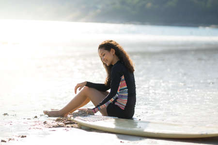 Curly blond long hair asian teenager girl with black surfing wet suit, sitting on sand beach beside surfboard. blue sea and island are backgroundの写真素材