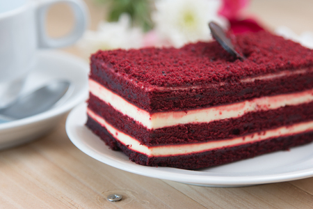 Red velvet cheese cake topping by dark chocolate on white plate and fork. Cup of coffee and vase of bouquet roses put on wooden table.の写真素材