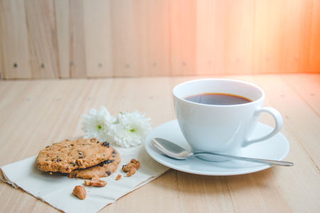 Cup of coffee and spoon on white plate, with almond chocolate ship cookies put near flower on wooden table background.の写真素材