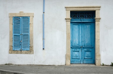Photo of typical Cypriot old aged white building with bright blue wooden door ,window with closed shutters and rainwater pipeの写真素材