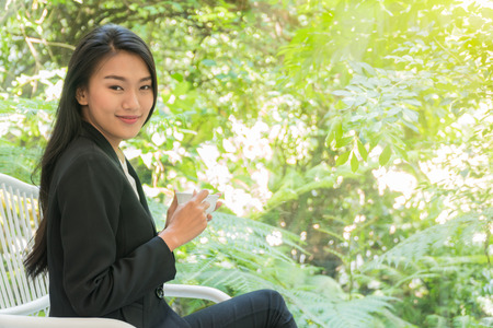 Beautiful young woman sitting in a chair holding a white cup coffee in her hand in the living room with garden background.の写真素材