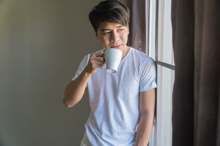 The handsome man stands at the window holding a white cup of coffee in his hand.の写真素材
