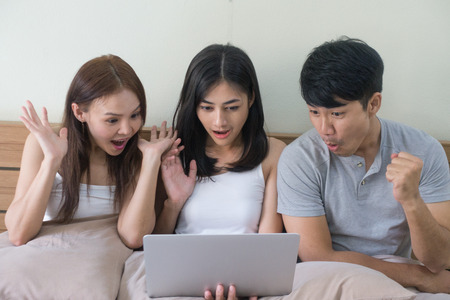 Three excited friends or roommates watching tv online in laptop sitting on bed.