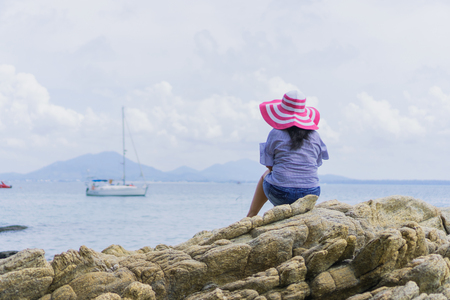 The back of the girl wearing a pink hat sitting on the rocks by the sea.の写真素材