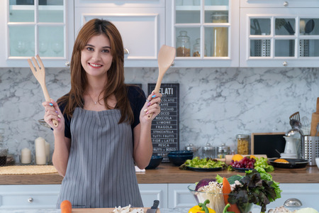 Cheerful young woman thinking about the menu while cooking in kitchen. Housewife holding wooden spoon in her hand. Healthy food conceptの写真素材